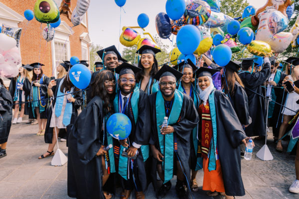 UVA graduates wearing caps and gowns are huddled together smiling at the camera. They hold blue balloons with the UVA Batten logo.