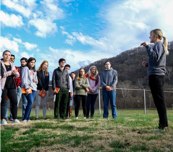 Students stand in a field with notebooks listening to professor talking